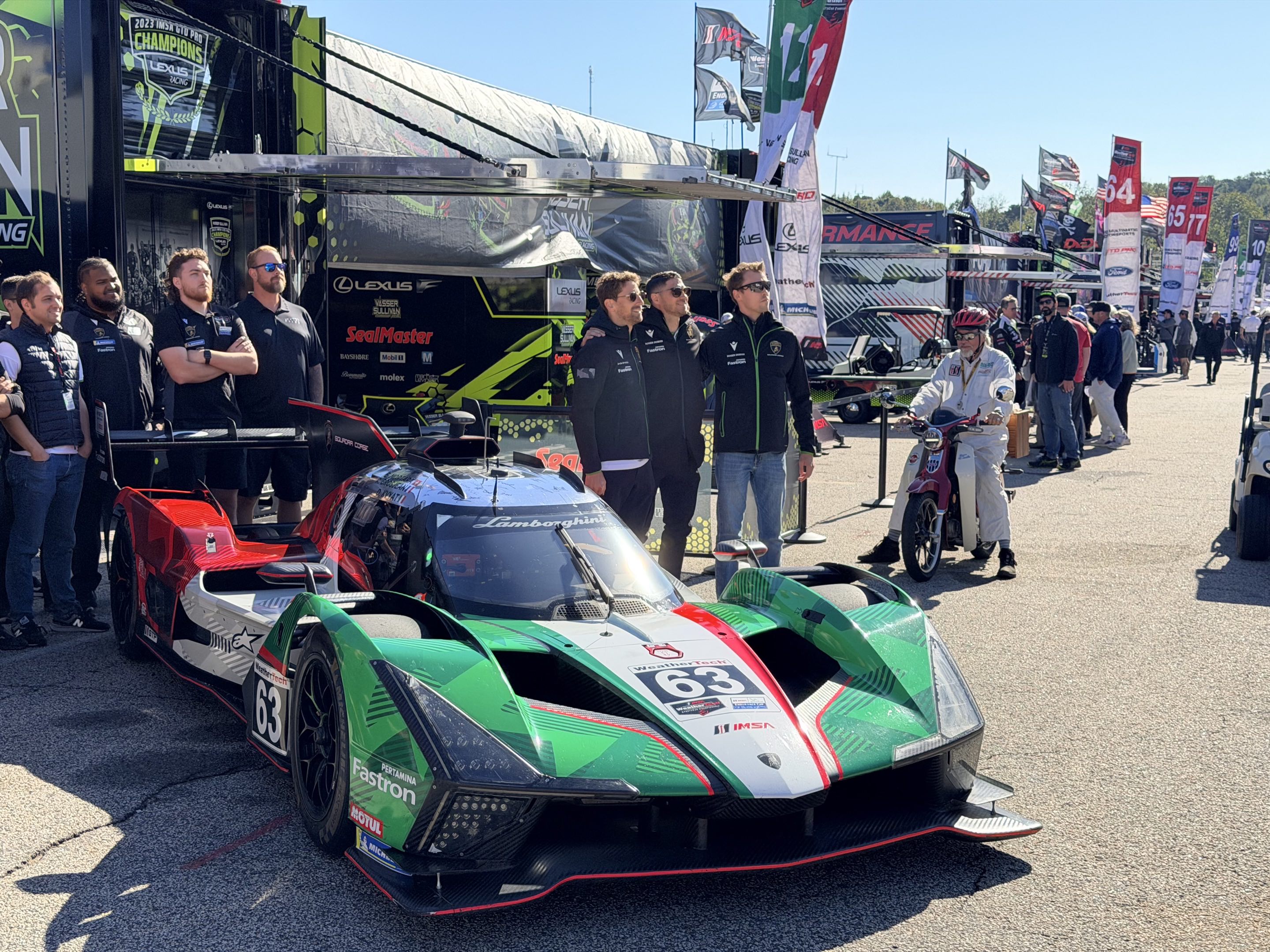 Three racing drivers in jackets and sunglasses standing next to a Lamborghini SC63 prototype race car